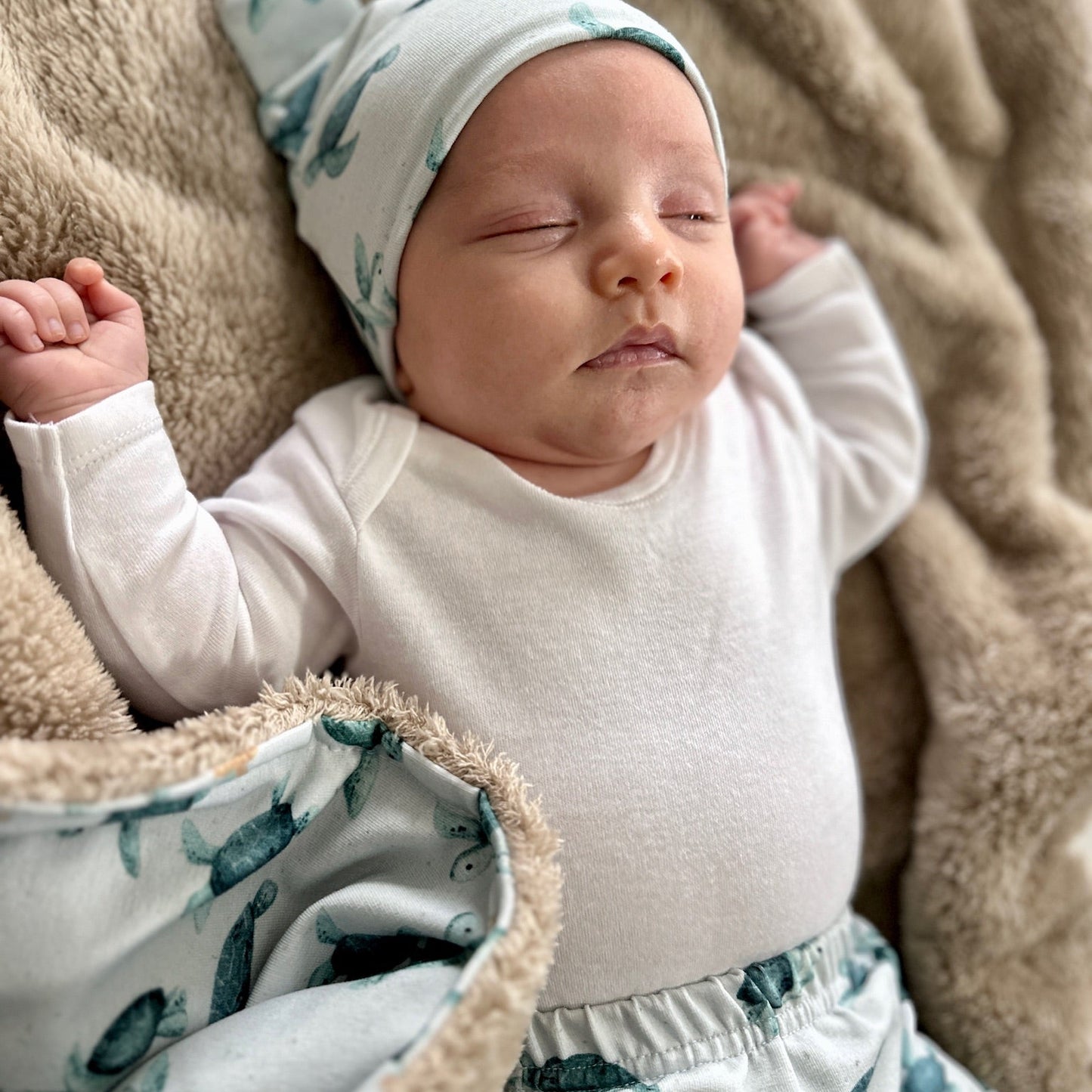 Newborn baby sleeping peacefully wearing a white outfit with green patterns on a soft brown blanket.