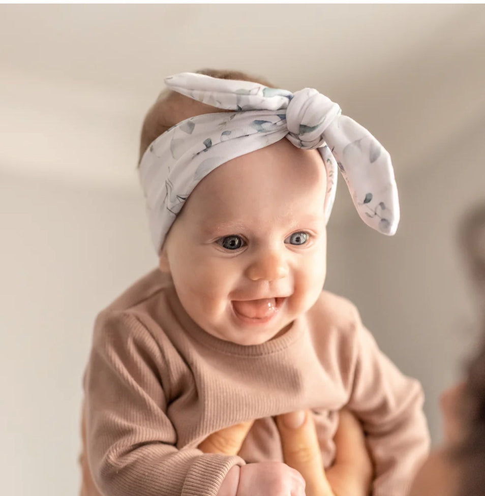 Baby wearing a white headband with a bow, sitting against a plain background